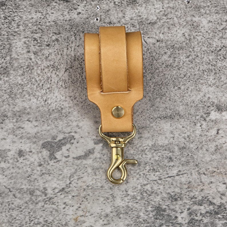 May include: A tan leather key fob with a brass-colored swivel clasp and a brass rivet. The key fob has a loop for attaching to a belt or bag. The leather is a light brown color.