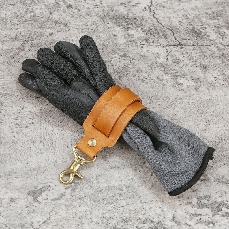 May include: Black work gloves with a textured surface, secured by a tan leather strap and a gold-colored clasp. The gloves are paired with gray knit sleeves.