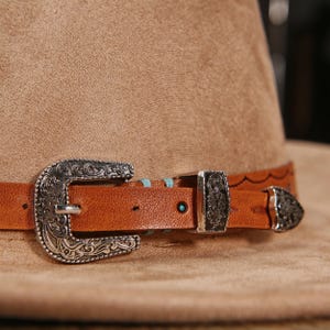 May include: A tan felt hat featuring a brown leather band adorned with an ornate silver-toned metal buckle, a matching metal keeper, and a pointed metal tip. The leather strap is accented with small turquoise-colored circular inlays.