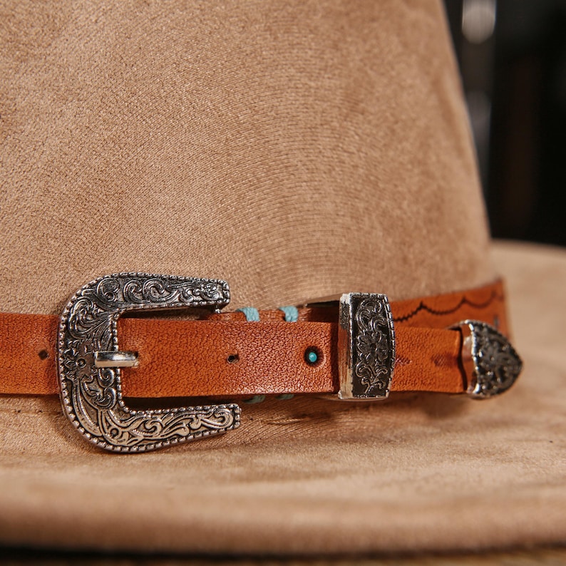 May include: A tan felt hat featuring a brown leather band adorned with an ornate silver-toned metal buckle, a matching metal keeper, and a pointed metal tip. The leather strap is accented with small turquoise-colored circular inlays.