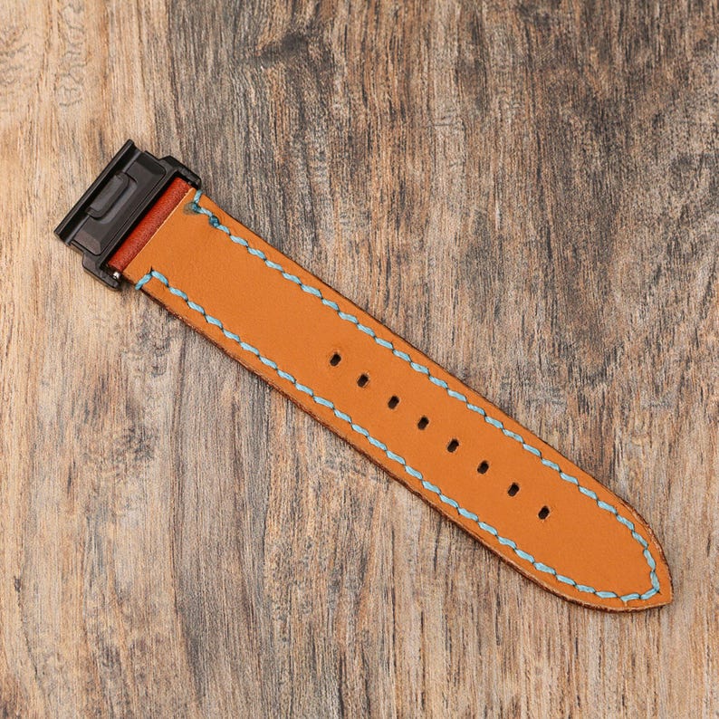 May include: A tan leather watch strap with light blue stitching and a black clasp. The strap has several holes for adjustment and is set against a wooden background.