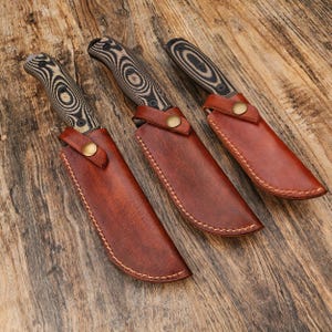 Puede incluir: Tres cuchillos de caza con mangos de madera y fundas de cuero. Los mangos de los cuchillos tienen un patr&oacute;n de remolino negro y marr&oacute;n. Las fundas son de cuero marr&oacute;n con cierre de bot&oacute;n de lat&oacute;n. Los cuchillos est&aacute;n sobre una superficie de madera.