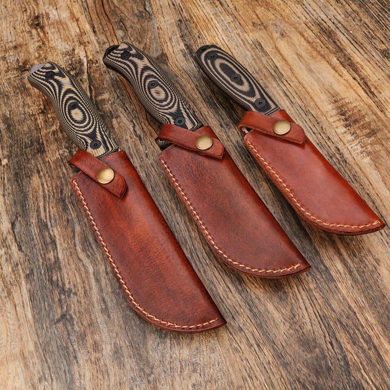 Puede incluir: Tres cuchillos de caza con mangos de madera y fundas de cuero. Los mangos de los cuchillos tienen un patr&oacute;n de remolino negro y marr&oacute;n. Las fundas son de cuero marr&oacute;n con cierre de bot&oacute;n de lat&oacute;n. Los cuchillos est&aacute;n sobre una superficie de madera.