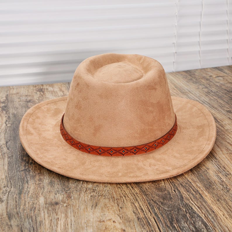 May include: A tan fedora hat with a flat crown and a wide brim. The hat features a textured, suede-like finish and is accented with a thin, dark orange band around the base of the crown, decorated with a repeating geometric pattern in black and teal.