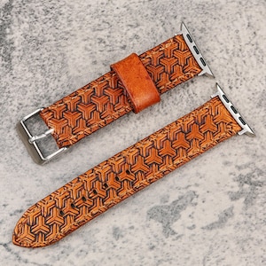 May include: A brown leather watch band with a geometric pattern. The band has a silver buckle and connectors. The leather is a warm brown color with a darker brown geometric design. The watch band is shown on a gray surface.