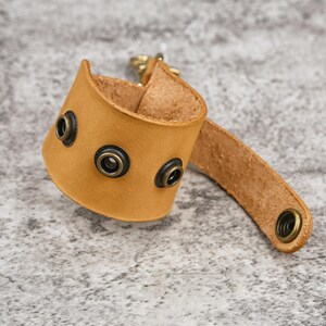 May include: A tan leather wrist cuff with three dark metal rivets and a matching strap. The cuff has a curved design and a metal clasp. The leather has a natural, textured appearance.