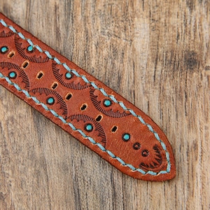 May include: Close-up of a brown leather watch strap with turquoise stitching and decorative circular patterns. The strap has several holes and is set against a wooden background.
