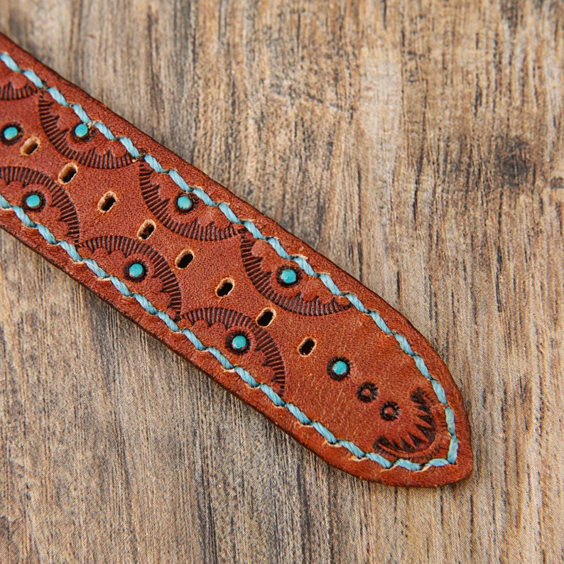 May include: Close-up of a brown leather watch strap with turquoise stitching and decorative circular patterns. The strap has several holes and is set against a wooden background.