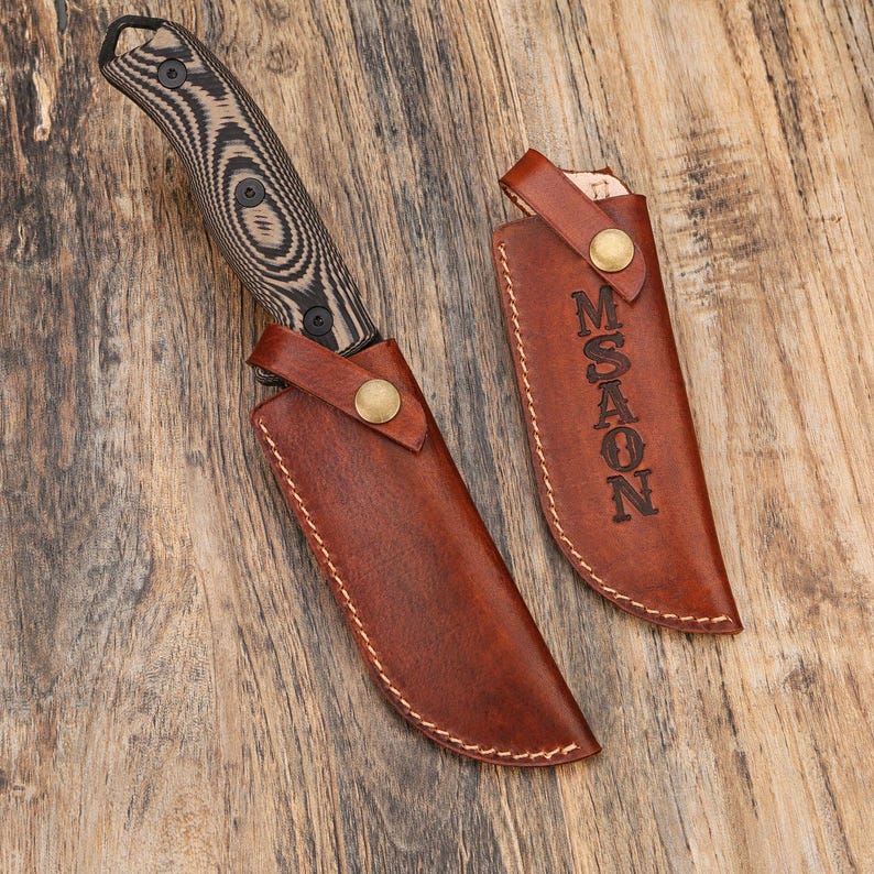 Puede incluir: Un cuchillo de hoja fija con mango negro y veteado de madera, junto con una funda de cuero marr&oacute;n. La funda se asegura con un bot&oacute;n de lat&oacute;n y tiene la palabra "MASON" grabada. El cuchillo y la funda se muestran sobre una superficie de madera.