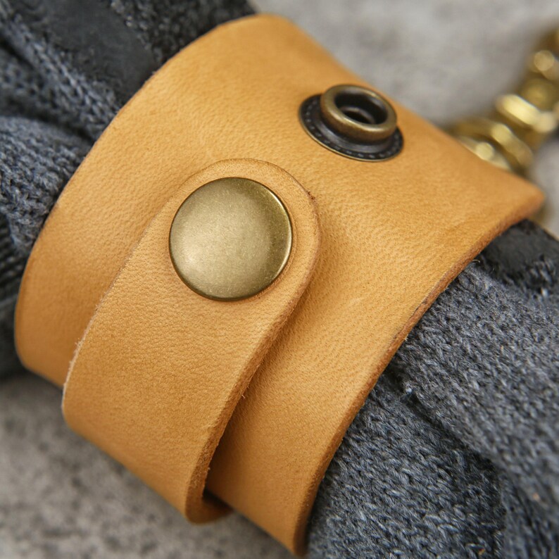 May include: A tan leather cuff with a brass snap closure, wrapped around a gray knitted item. The cuff has a simple design with a rounded edge and a contrasting dark metal rivet.