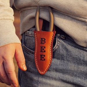 May include: A brown leather tool holder with the letters "BEE" embossed on the front, stitched with white thread. The holder contains a pair of gray gardening shears. The holder is tucked into a blue jeans pocket.