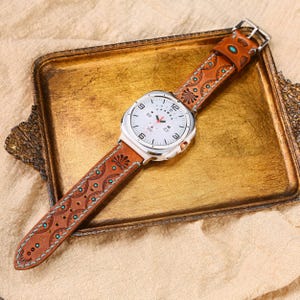 May include: A silver-toned smartwatch with a white face and a brown leather band. The band features a detailed embossed pattern with turquoise accents. The watch is displayed on a gold-colored tray.