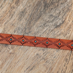 May include: A tan leather belt strap featuring a repeating embossed geometric pattern with small, circular turquoise-colored inlays centered within each diamond-shaped motif along the length of the material.