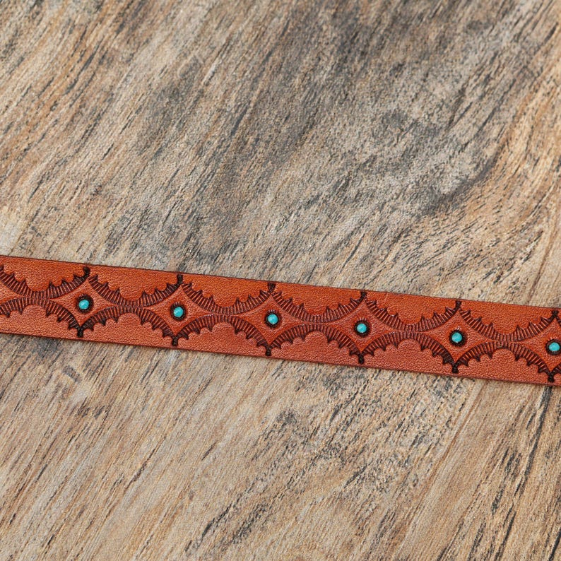 May include: A tan leather belt strap featuring a repeating embossed geometric pattern with small, circular turquoise-colored inlays centered within each diamond-shaped motif along the length of the material.