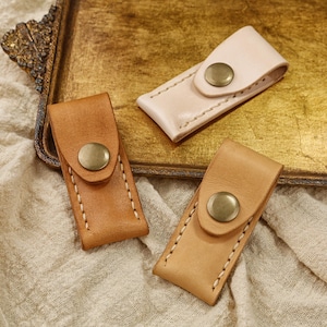 May include: Three handcrafted leather sheaths in varying shades of brown and beige. Each sheath features a brass button closure and white stitching. The sheaths are displayed on a gold-toned tray and a beige fabric.