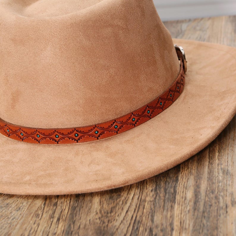 May include: A tan fedora hat with a smooth, suede-like texture. The hat features a brown leather band decorated with a repeating geometric pattern and small turquoise-colored circular accents. A metallic buckle is visible on the side of the band.