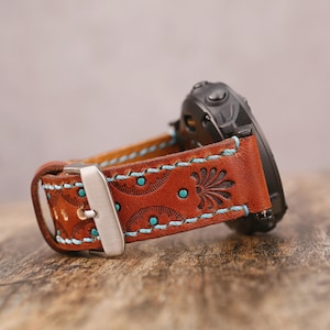 May include: Brown leather watch band with turquoise stitching and accents. The band features a floral design and a silver buckle. A black watch is attached to the band.