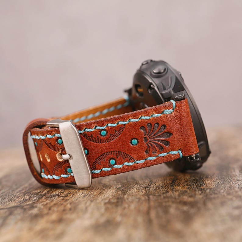 May include: Brown leather watch band with turquoise stitching and accents. The band features a floral design and a silver buckle. A black watch is attached to the band.