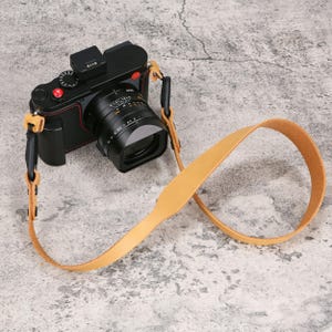 May include: A black digital camera with a prominent lens and a red shutter button, attached to a tan leather camera strap. The strap features black metal hardware connectors on both sides, securing the light brown leather band to the camera body.