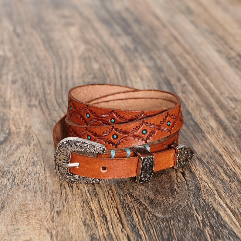 May include: A tan leather belt featuring an intricate embossed pattern with small turquoise circular inlays. The belt includes a silver-toned metal buckle with ornate floral engravings, a matching metal keeper, and a pointed metal tip.