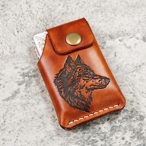 May include: A brown leather card holder with a detailed black wolf head design. The card holder has a flap with a brass button closure and white stitching along the bottom edge. The card holder is designed to hold cards.