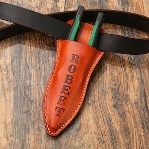 May include: A brown leather knife sheath with the name "ROBERT" embossed vertically. Two green and black handled tools are inserted into the sheath. The sheath is attached to a black leather belt. The background is a wooden surface.