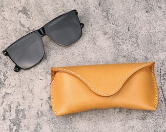 May include: Black sunglasses with a rectangular frame next to a tan leather glasses case. The case is stitched with white thread and has a curved top. The sunglasses have dark lenses and a black frame.