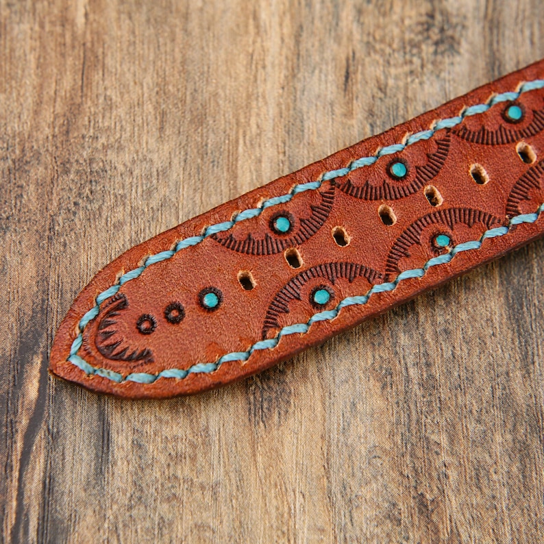 May include: Close-up of a brown leather watch strap with turquoise stitching and accents. The strap features a detailed embossed pattern and several small holes for adjustment. The edges are stitched with turquoise thread.