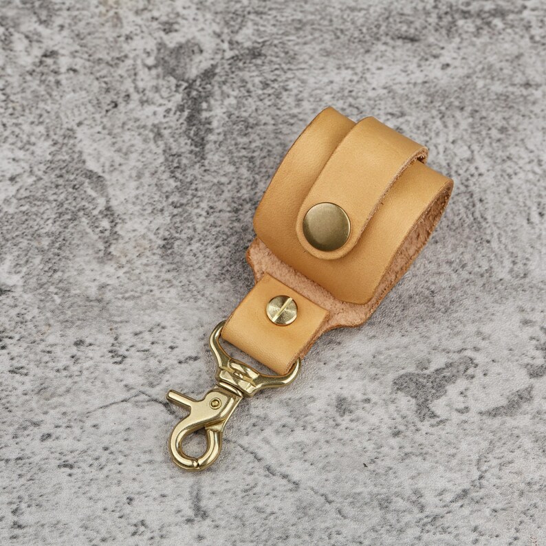May include: A light brown leather keychain holder with a brass clasp and button closure. The holder is made of two overlapping pieces of leather, with a brass button on the front. The clasp is a standard swivel type.