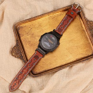May include: A black smartwatch with a brown leather band featuring a decorative floral and dot pattern with turquoise accents. The watch face displays data, and the band is secured with a silver buckle. The watch is on a gold tray.