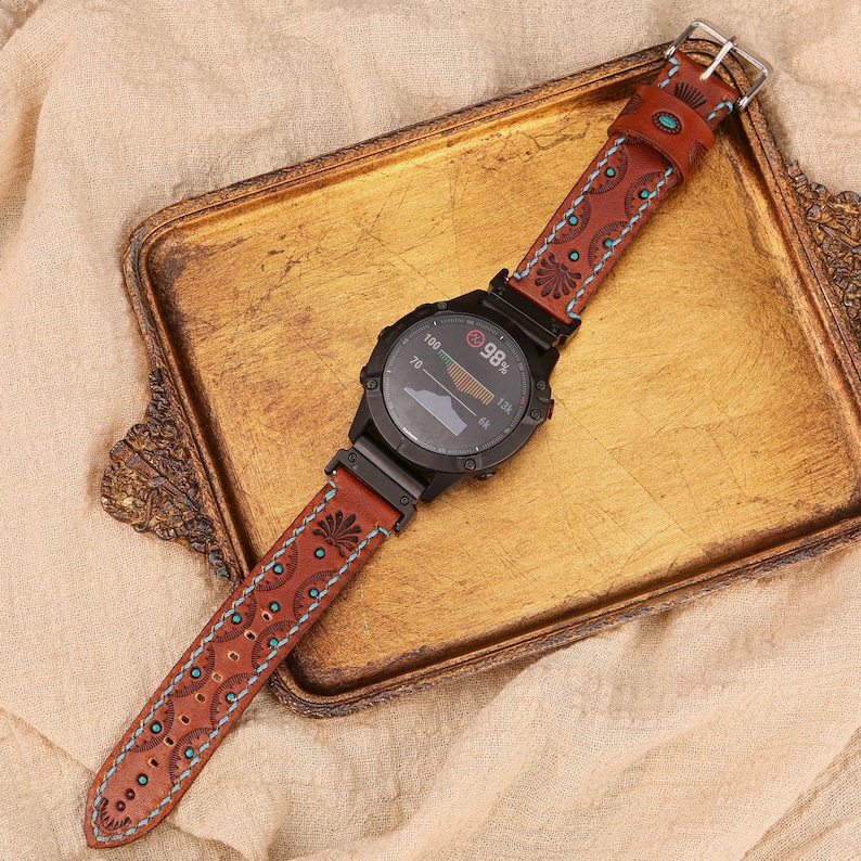 May include: A black smartwatch with a brown leather band featuring a decorative floral and dot pattern with turquoise accents. The watch face displays data, and the band is secured with a silver buckle. The watch is on a gold tray.