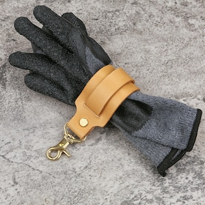 May include: A pair of work gloves with black textured palms and gray knit backs, secured by a tan leather strap and a gold-colored clasp. The gloves are designed for protection and are likely used for gardening or construction.
