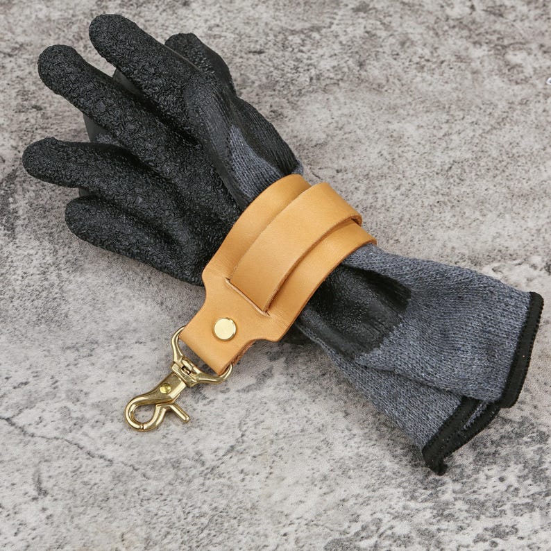 May include: A pair of work gloves with black textured palms and gray knit backs, secured by a tan leather strap and a gold-colored clasp. The gloves are designed for protection and are likely used for gardening or construction.