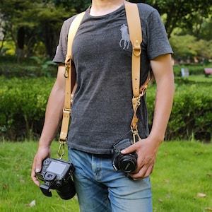 May include: A tan leather camera harness with gold-colored metal hardware. The harness is designed to hold two cameras, with each camera attached to a hook. The person in the image is wearing a gray t-shirt and blue jeans.