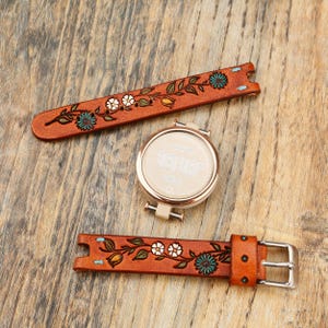 May include: A round smartwatch with a rose gold bezel and a digital display, accompanied by two detachable tan leather watch straps. The straps feature embossed floral patterns with hand-painted teal and white details and light blue stitching.