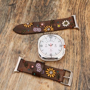 May include: A silver smartwatch with a white digital face featuring a circular display, accompanied by two dark brown leather watch straps. The straps are decorated with embossed and painted yellow, white, and pink flower and leaf patterns.