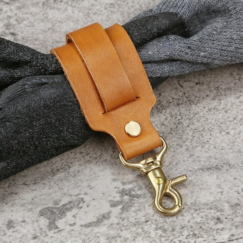 May include: A tan leather strap with a brass-colored clasp and rivet. The strap is looped over a dark gray object, possibly a handle or strap. The clasp is open, ready to attach to another item.