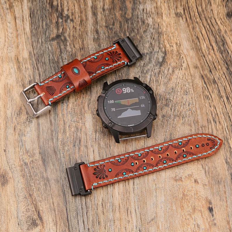 May include: A black smartwatch with a round face and two brown leather watch bands. The bands are decorated with turquoise accents and light blue stitching. The watch face displays a percentage and a graph.