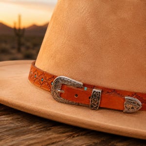 May include: A tan felt hat featuring a brown leather band with intricate embossed patterns, small turquoise stone accents, and a silver-toned metal buckle set with ornate floral engravings. The hat has a wide brim and a structured crown.