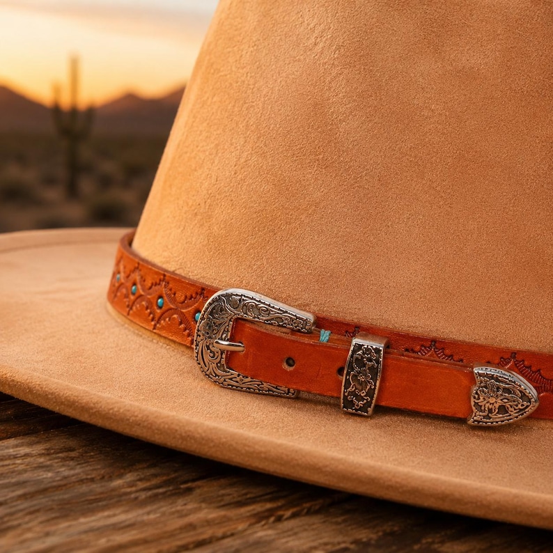 May include: A tan felt hat featuring a brown leather band with intricate embossed patterns, small turquoise stone accents, and a silver-toned metal buckle set with ornate floral engravings. The hat has a wide brim and a structured crown.