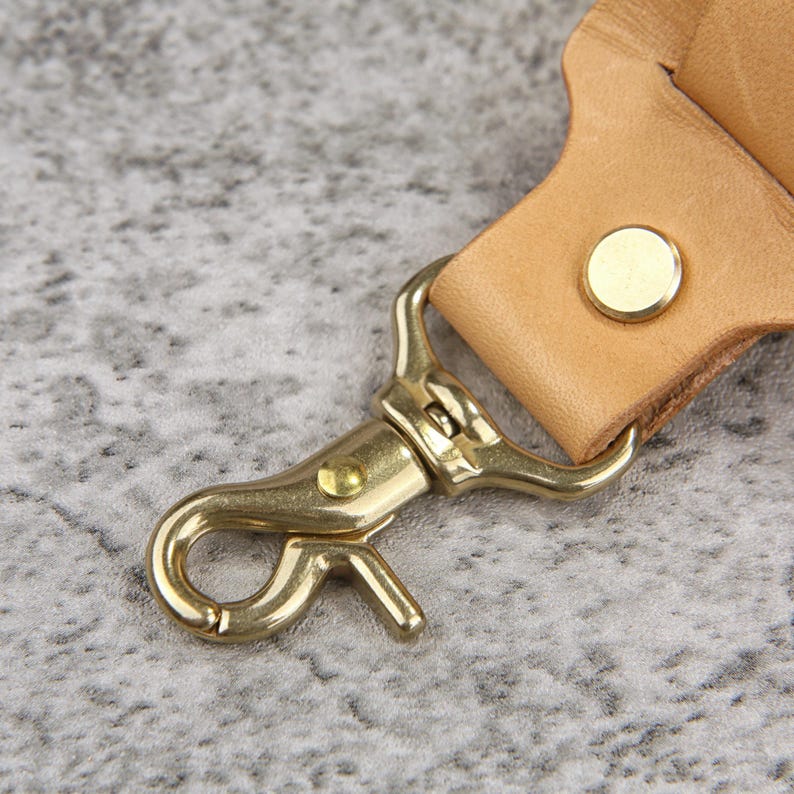 May include: Close-up of a brass-colored swivel clasp attached to a light brown leather strap. The clasp is open, revealing its hook and lever mechanism. A brass rivet secures the leather. The background is a mottled gray.