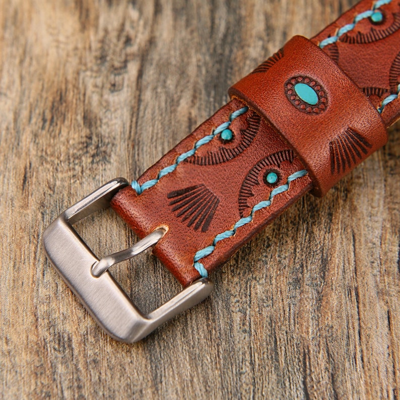 May include: Close-up of a brown leather watch strap with a silver buckle. The strap features embossed geometric patterns, turquoise accents, and light blue stitching. The watch band is on a wooden surface.