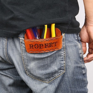 May include: A brown leather tool holder with the name "ROBERT" embossed on the front, visible in the back pocket of blue jeans. The holder contains several tools with red, blue, black, and yellow handles. The holder has a decorative scalloped edge.