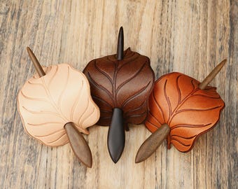 May include: Three leather leaf-shaped hair pins in varying shades of brown and tan. Each pin features a wooden stick and a dark, elongated base. The pins are arranged on a wooden surface.
