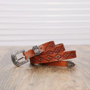 May include: A tan leather belt featuring intricate embossed patterns, turquoise-colored accents, and silver-toned metal hardware including a decorative buckle, a matching metal keeper, and a pointed metal tip at the end of the strap.