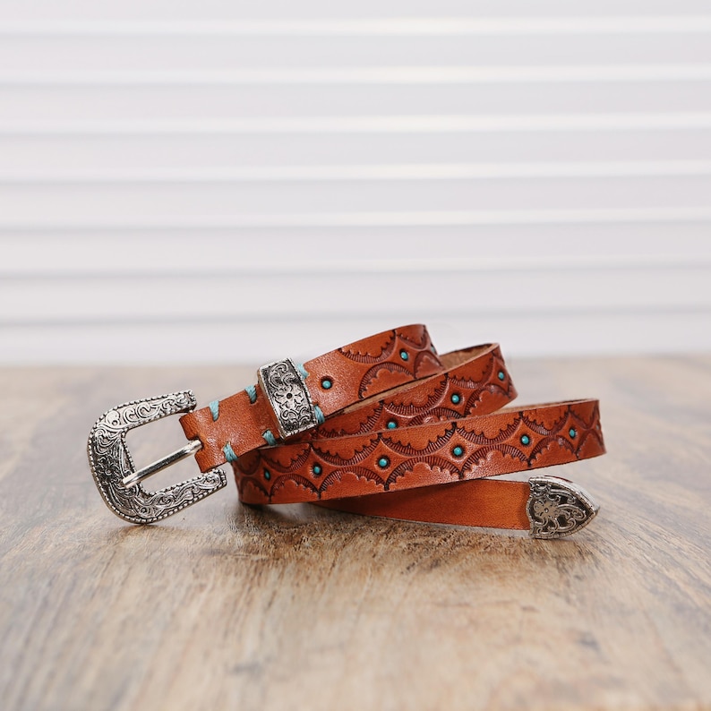 May include: A tan leather belt featuring intricate embossed patterns, turquoise-colored accents, and silver-toned metal hardware including a decorative buckle, a matching metal keeper, and a pointed metal tip at the end of the strap.