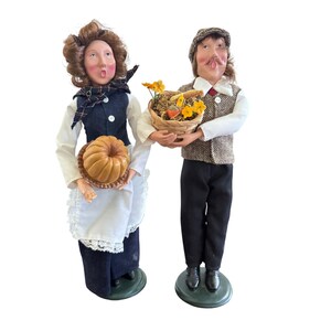 May include: A pair of decorative figurines depicting a man and woman. The woman holds a pumpkin on a plate, wearing a white blouse, blue vest, and skirt. The man carries a basket of flowers and produce, dressed in a tweed vest, cap, and black pants.
