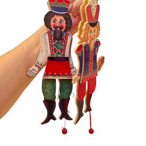 May include: Two wooden, articulated figures of men in traditional costumes. One figure wears a red and white outfit with a black beard and mustache. The other figure wears a red and blue outfit with a blonde beard. Both figures have red strings and wooden beads.
