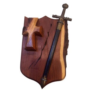 May include: A wooden shield with a carved cross and a decorative sword. The shield is made of dark brown wood with a natural edge. The sword has a bronze-colored hilt and a black scabbard.