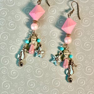 Könnte beinhalten: Ein Paar Ohrhänger mit Meeresmotiven. Jeder Ohrring hat eine rosa geometrische Perle, eine rosa Perle und Charms, darunter ein Seepferdchen, einen Seestern und einen Fisch. Türkisfarbene Perlen sorgen für Farbe. Die Ohrringe haben bronzefarbene Ohrhaken.